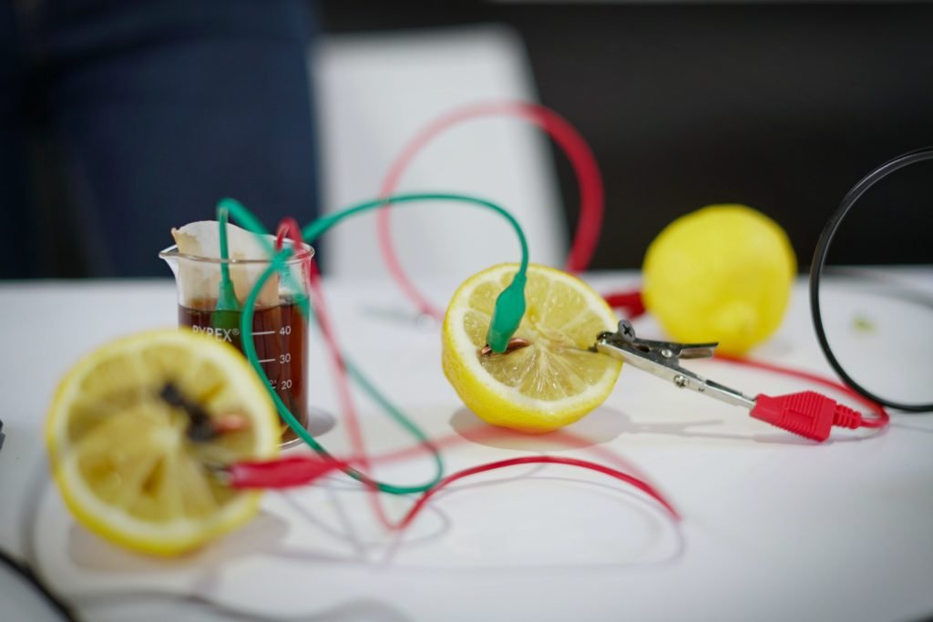 A simple scientific experiment setup showing a lemon battery with wires and alligator clips attached to a cut lemon, converting chemical energy into electrical energy.